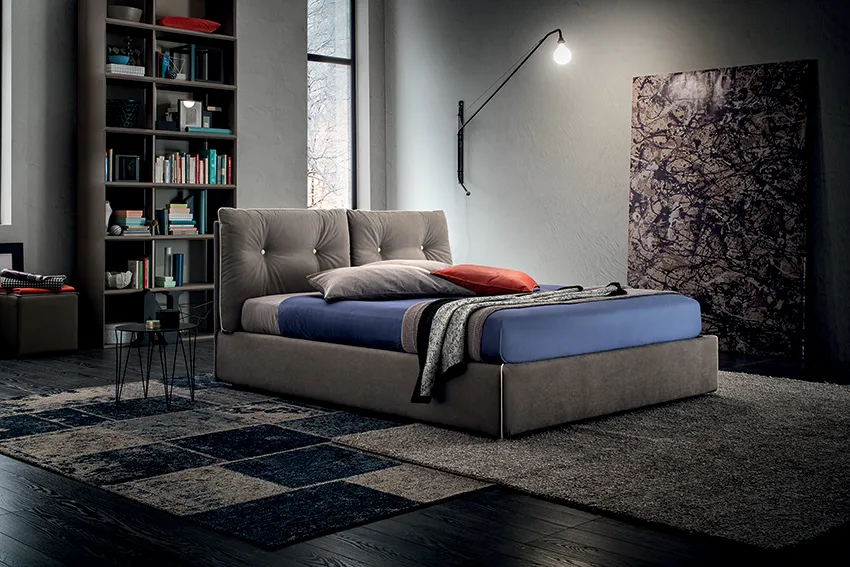 Camera da letto dal mood industriale con letto imbottito grigio, biancheria blu e cuscino rosso, libreria e lampada da parete orientabile