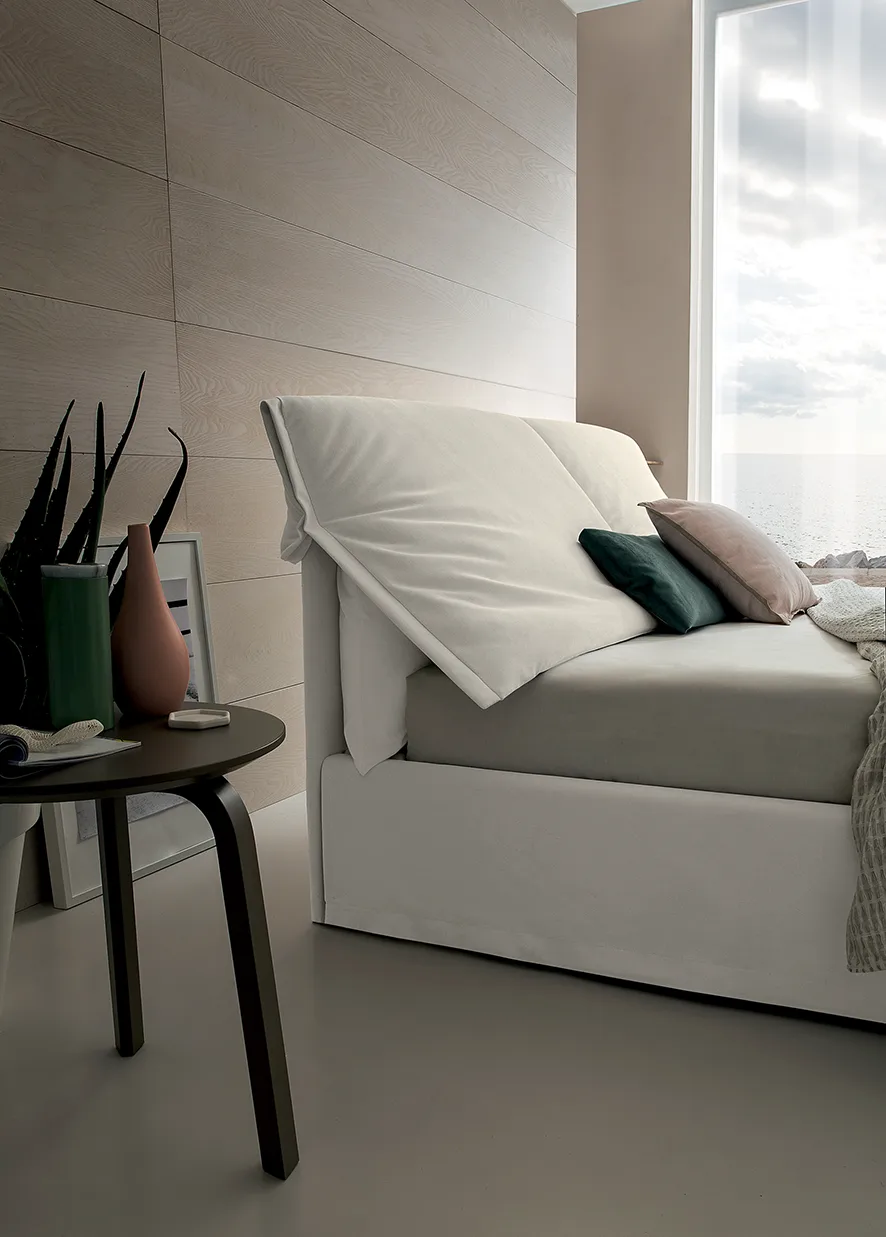 Camera da letto luminosa con letto imbottito bianco dalla testiera morbida, comodino rotondo e vista mare dalla finestra