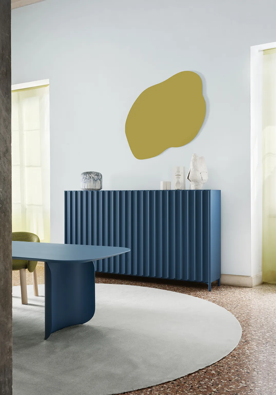 Ambiente con credenza blu a doghe verticali, tavolo sagomato blu, elemento decorativo a parete organico color oliva e tappeto chiaro