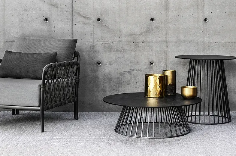 Poltrona outdoor con intreccio in corda su struttura metallica, tavolini rotondi in metallo nero e portacandele dorati su sfondo in cemento